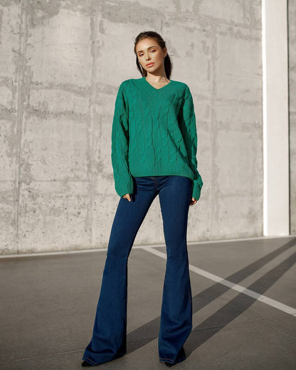 Woman wearing a green sweater and blue flared pants standing against a concrete wall.
