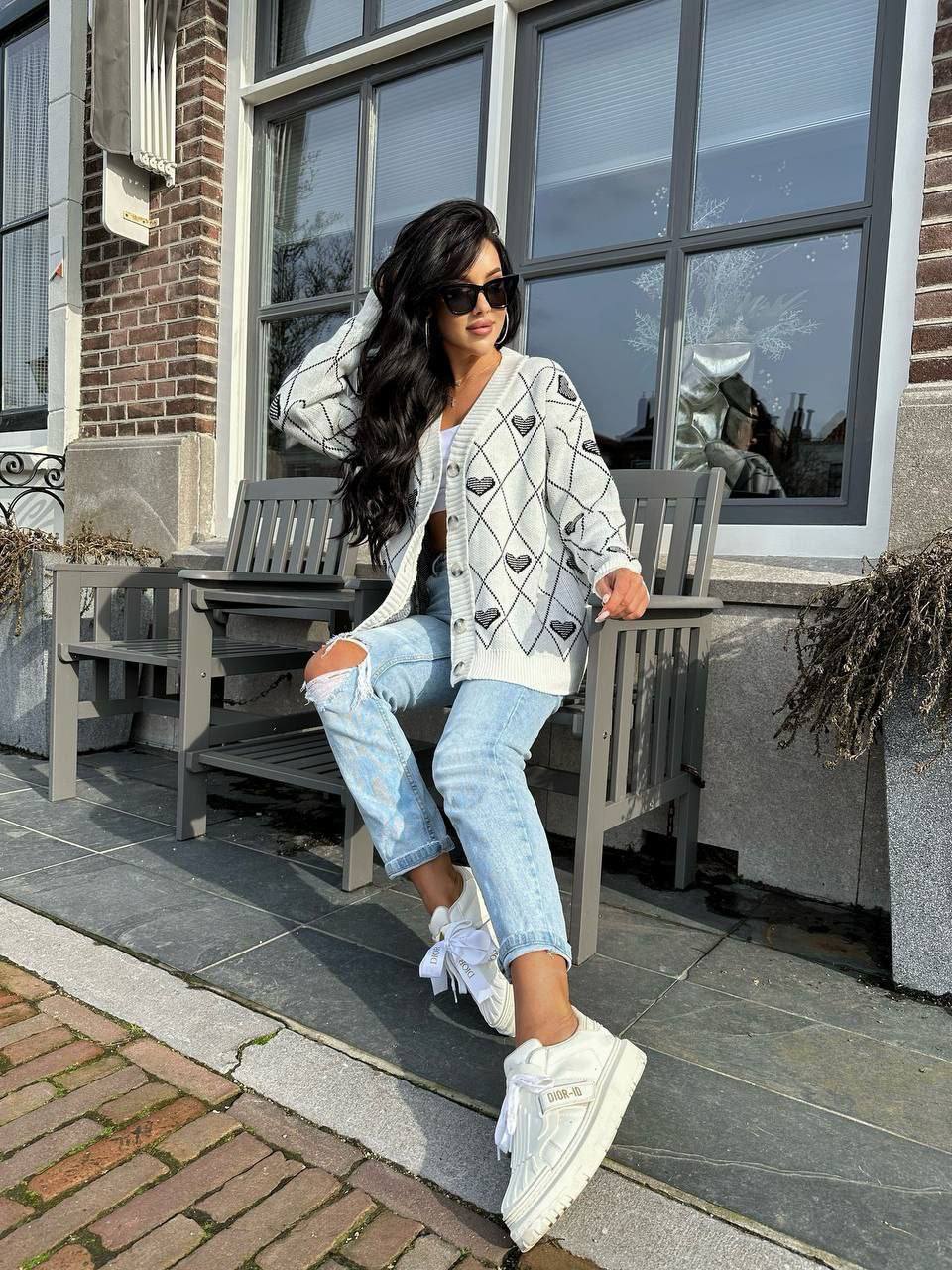 Woman sitting on a bench wearing a patterned cardigan, light blue jeans, and white sneakers.