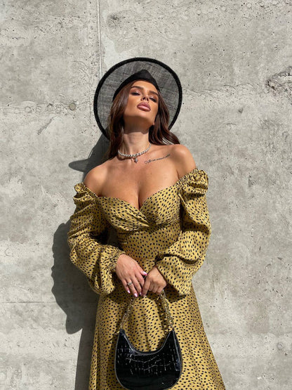 Woman in a yellow off-shoulder dress and black hat against a concrete wall.