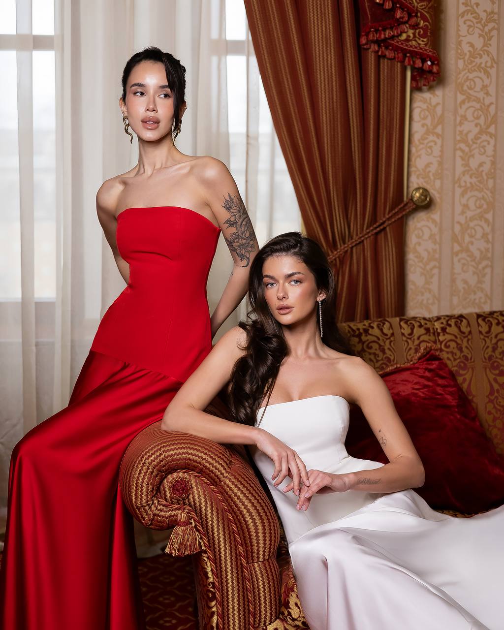 Two women in elegant dresses posing in a luxurious room with ornate decor.