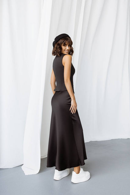 Woman wearing a black sleeveless top and long skirt standing against a white curtain.