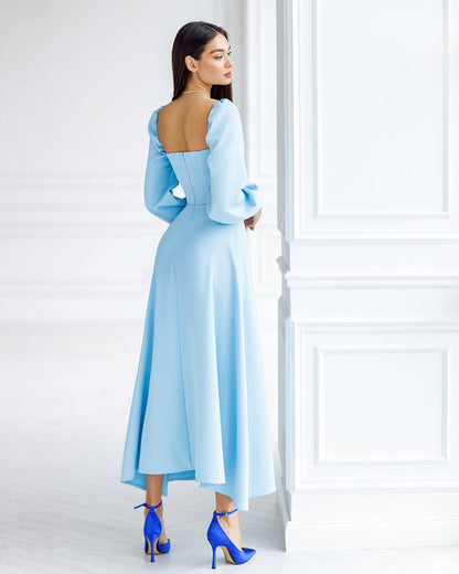 Woman wearing a light blue dress with long sleeves and a high-low hem, standing against a white wall.