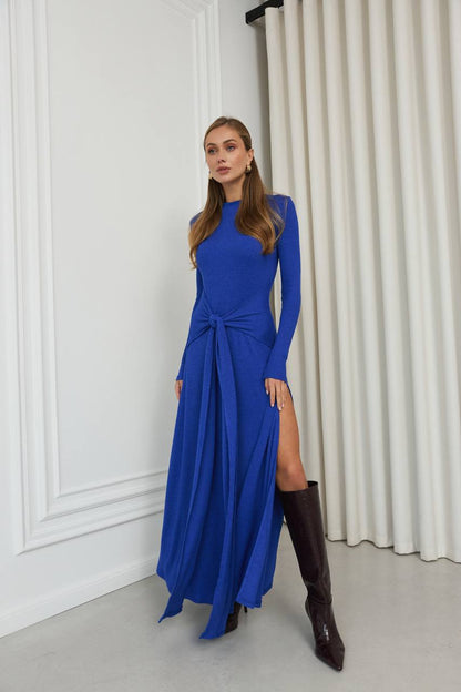 Woman wearing a blue dress with long sleeves and a slit, standing against a white wall.