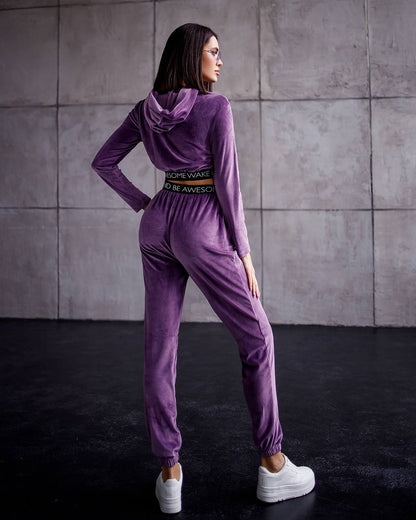 Woman wearing a purple tracksuit with a belt against a gray tiled wall.