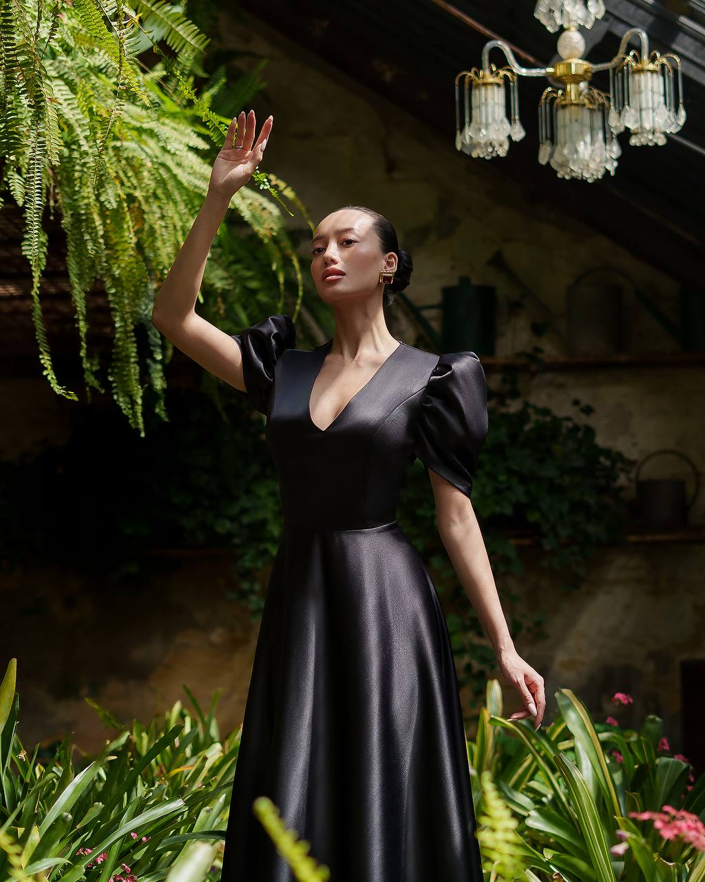 Woman in a black dress standing among plants with a chandelier in the background