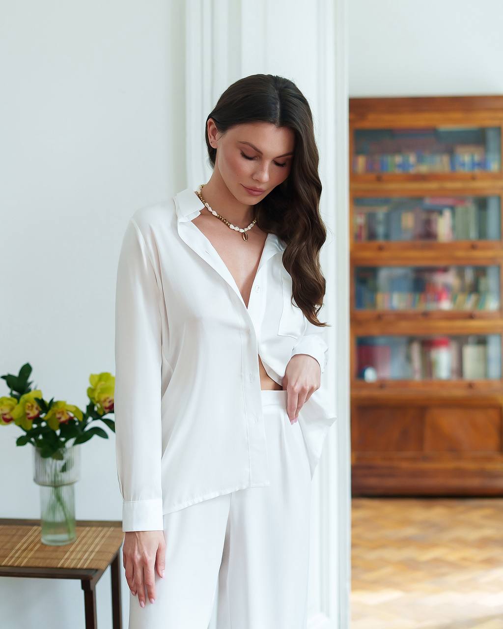 Woman wearing a white shirt in a room with a bookshelf and flowers.