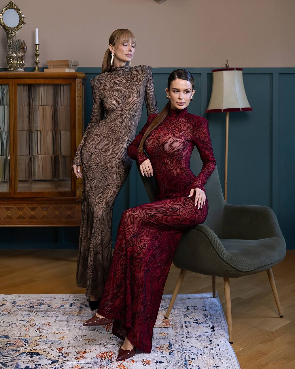 Two women in elegant dresses posing in a stylish room with a bookshelf and lamp.