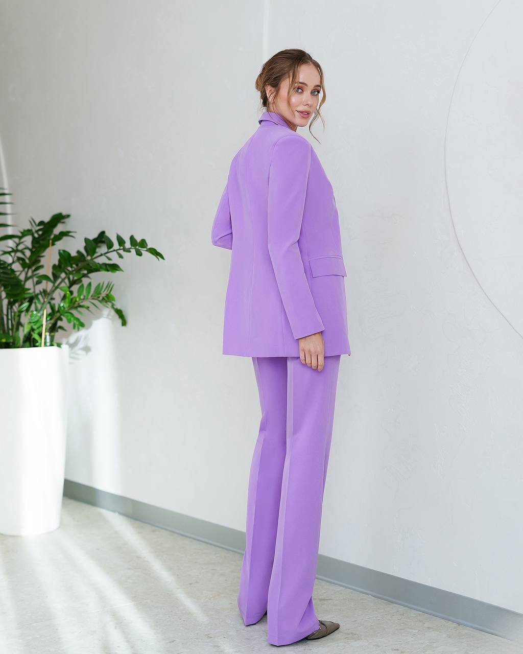 Woman wearing a purple suit standing in an indoor setting with a plant and white wall.