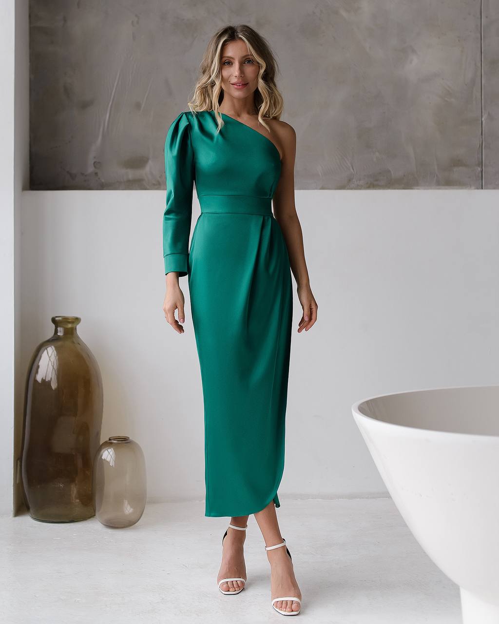 Woman wearing a green one-shoulder dress in a minimalistic setting with a vase and table.