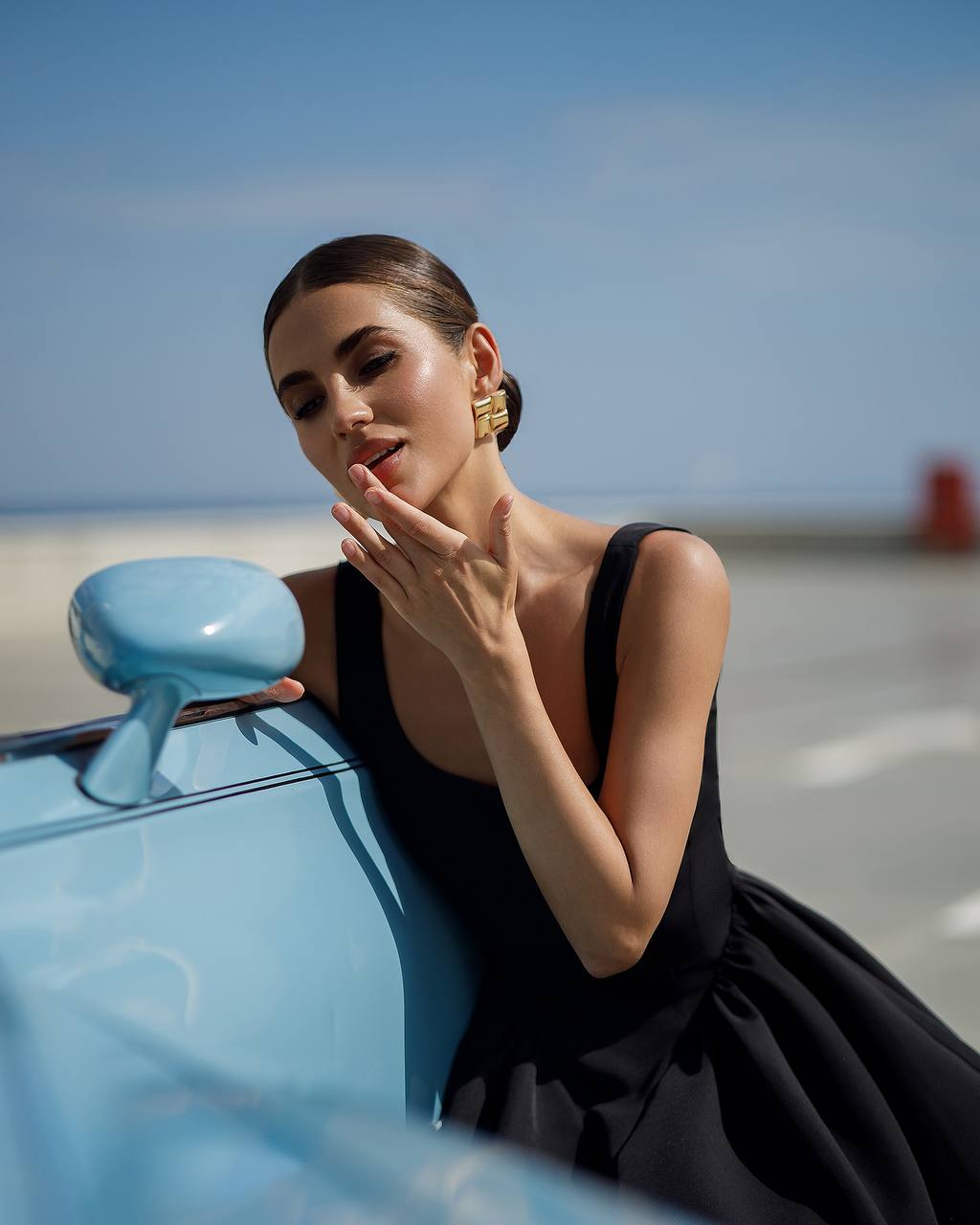 Woman in a black dress leaning against a blue car with a clear sky background