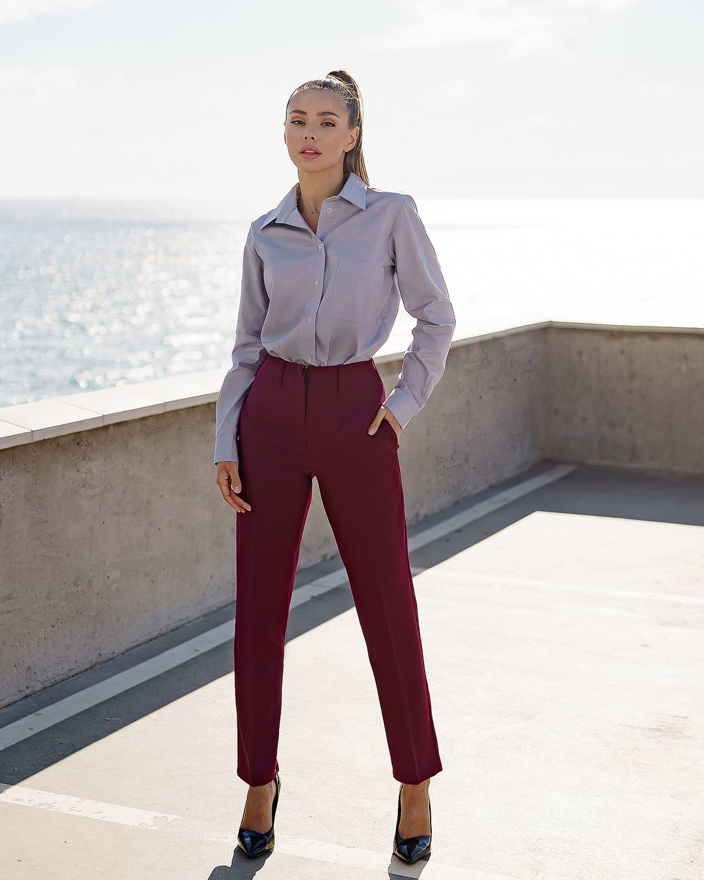 Woman in a light gray shirt and burgundy pants standing on a rooftop with water in the background