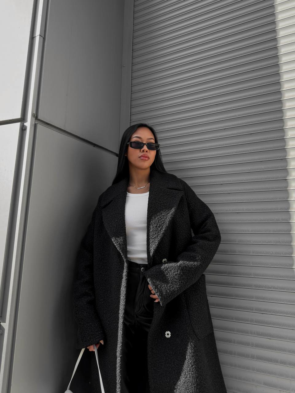 Woman wearing a black coat and sunglasses against a gray wall