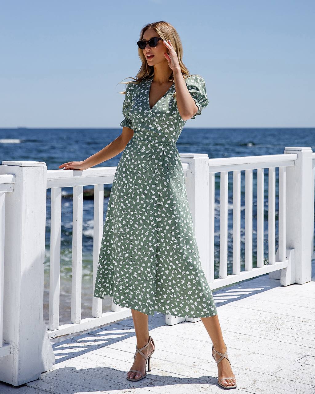 Woman in a green floral dress standing on a deck by the ocean.