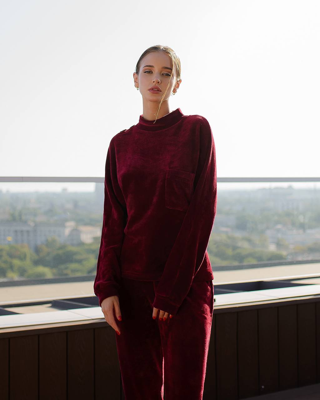 Person wearing a burgundy outfit standing in front of a window with a cityscape view.