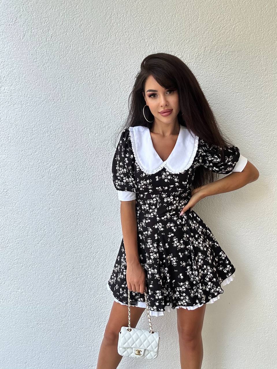 Woman wearing a black floral dress with white collar and cuffs, holding a white handbag against a light background.