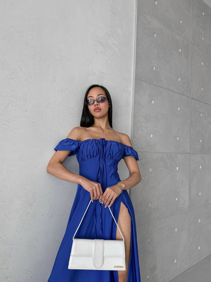 Woman in a blue off-shoulder dress holding a white handbag against a gray wall.