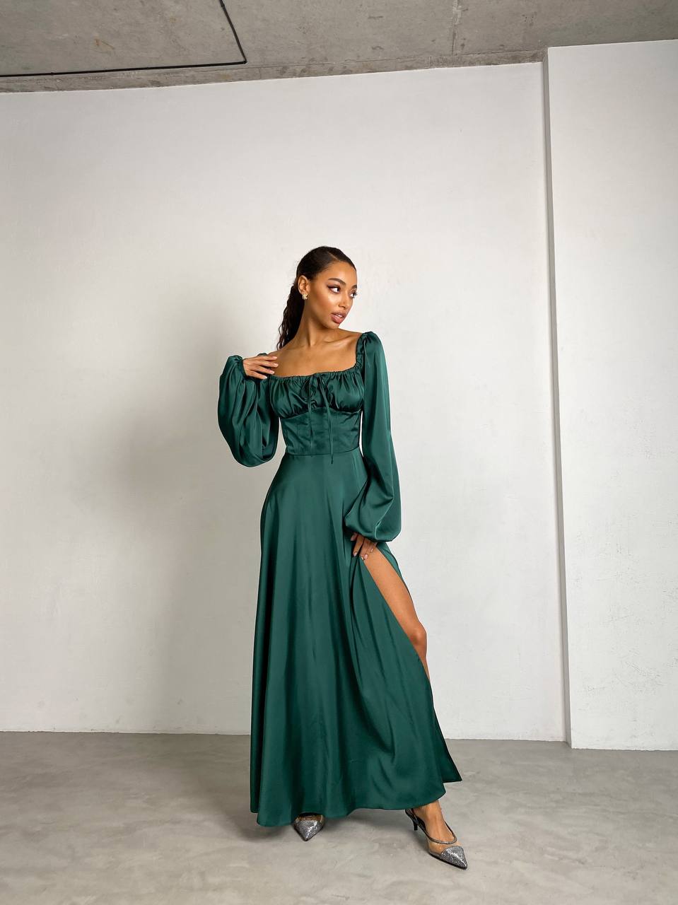 Woman wearing a green dress with long sleeves and a slit, standing against a plain white wall.