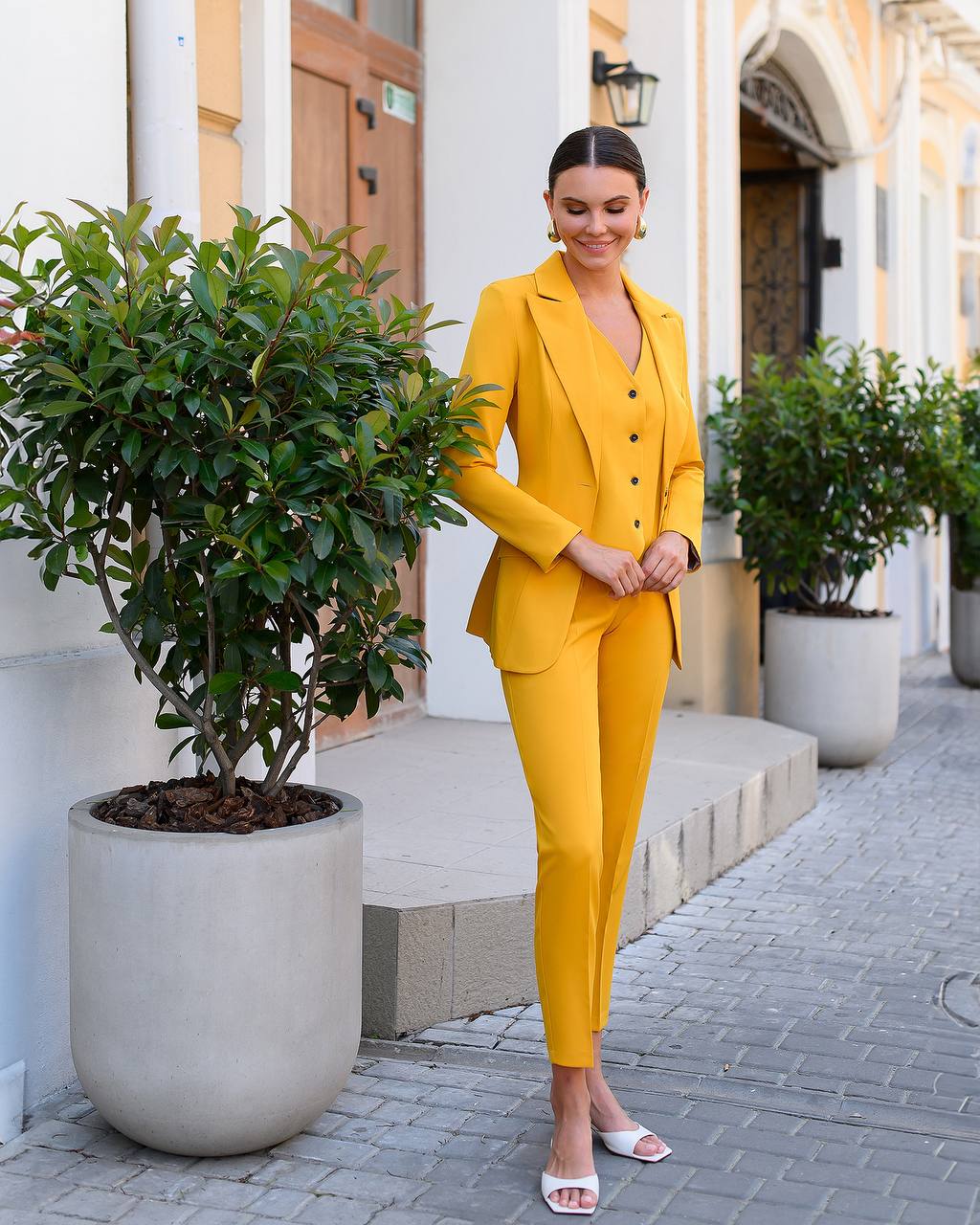 Woman in a yellow suit standing outdoors next to a building with plants.