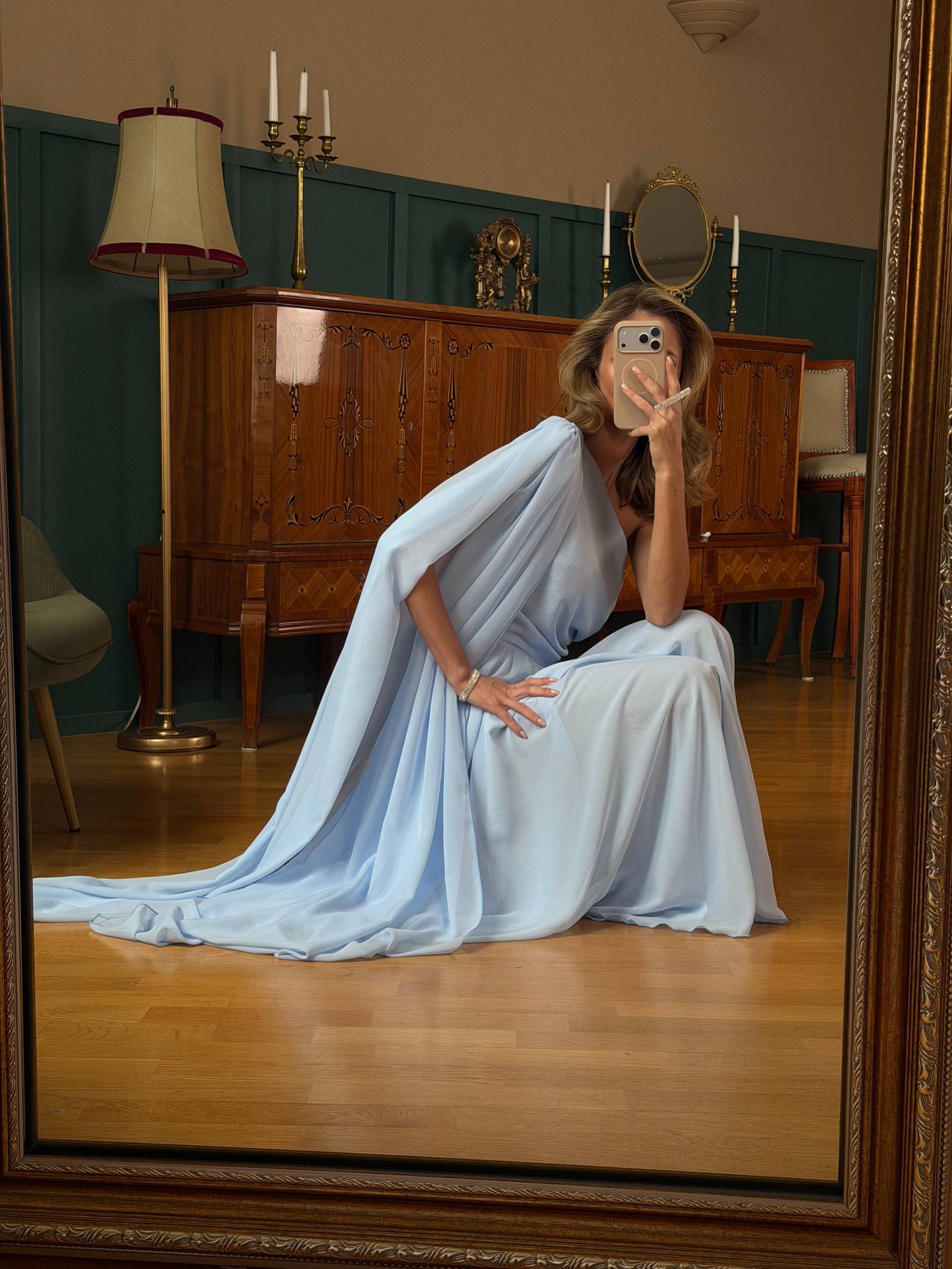 Woman in a light blue dress sitting on the floor in a room with wooden furniture and a mirror.
