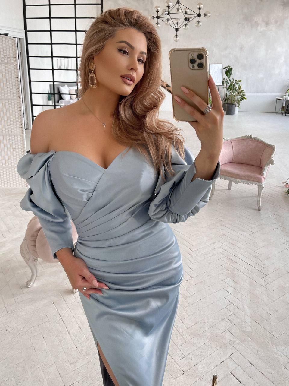 Woman in a light blue off-shoulder dress taking a mirror selfie in a stylish room.