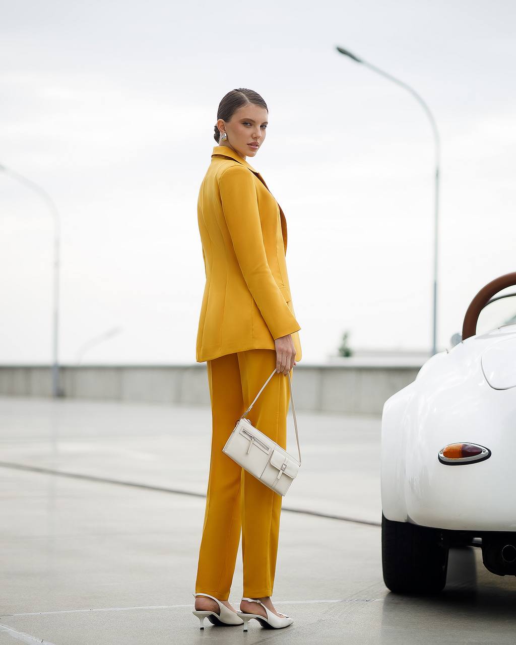 Woman in a mustard yellow suit standing next to a white car in an urban setting