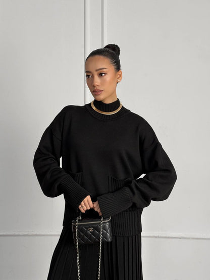 Woman wearing a black outfit holding a handbag against a plain background