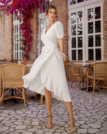 Woman in a white dress standing in an outdoor setting with wicker chairs and a brick building.