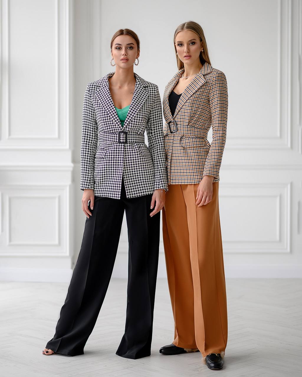 Two women wearing checkered blazers and wide-leg pants against a white wall.