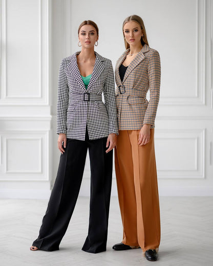 Two women wearing checkered blazers and wide-leg pants against a white wall.