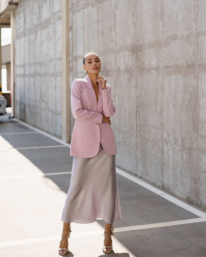 A woman stands in a concrete corridor wearing a light pink blazer, a pale pink satin midi skirt, and tan strappy high-heeled sandals.