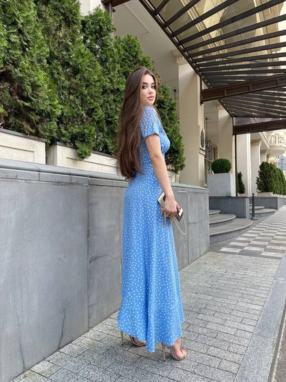 Woman in a blue dress standing on a sidewalk with greenery and a building in the background