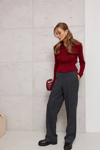 Woman wearing a red sweater and gray pants against a light gray wall.