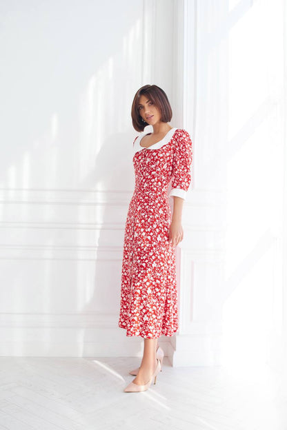 Woman wearing a red floral dress standing against a white curtain.