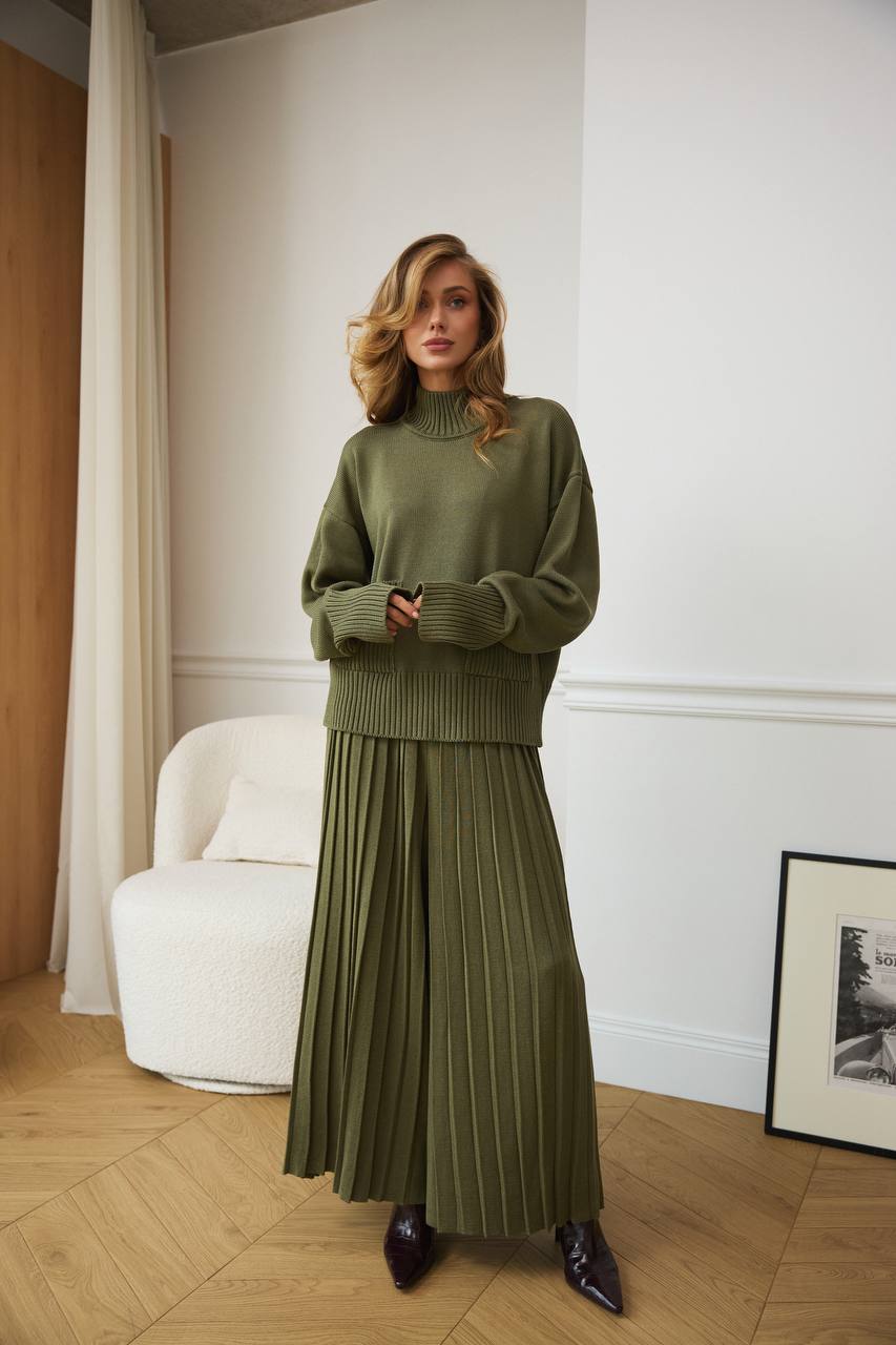 Woman wearing a green sweater and pleated skirt standing in a room with a white armchair and framed picture.