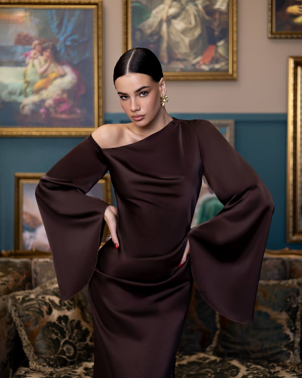 A woman modeling a dark brown off-the-shoulder satin dress with dramatic bell sleeves, posed in an elegant room with framed paintings on the wall.