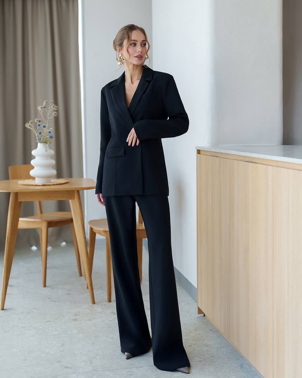 Woman in a black suit standing in a modern room with wooden furniture.