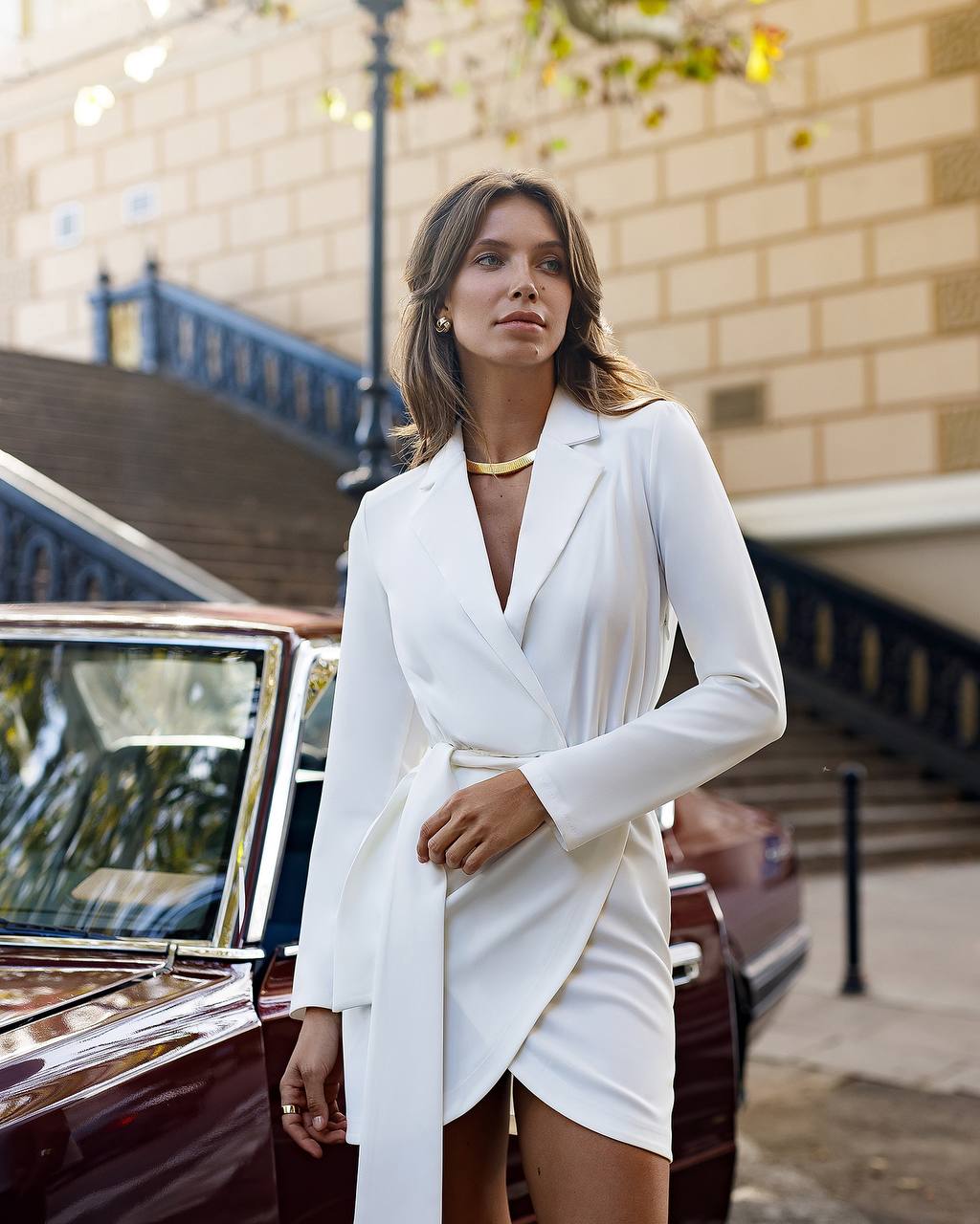 Woman in a white blazer dress standing next to a vintage car with a cityscape background