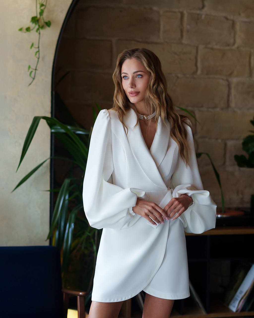 Woman wearing a white blazer dress in an indoor setting with plants and a brick wall.