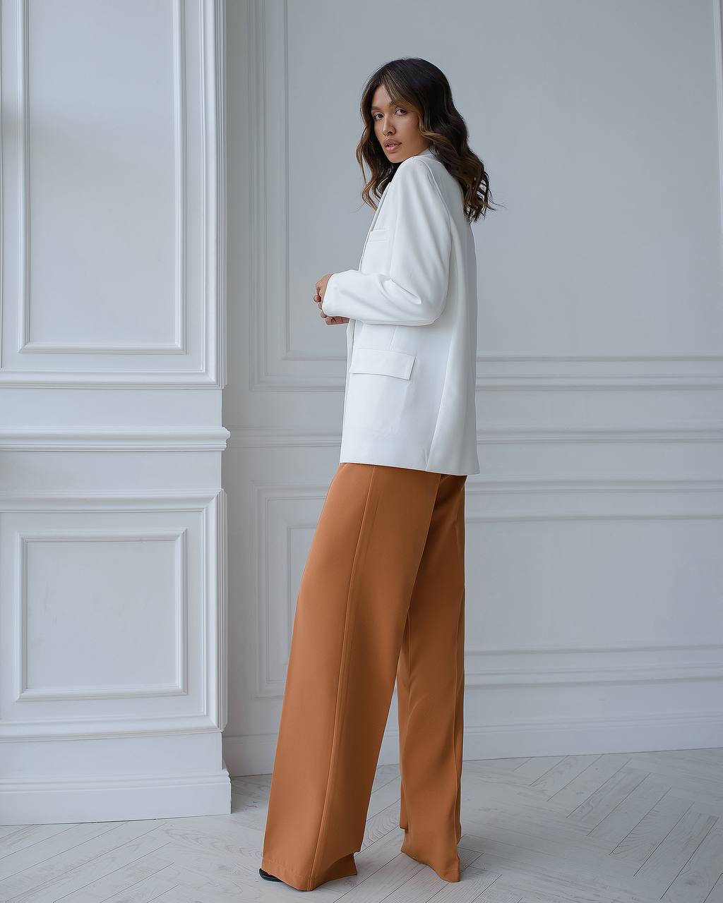 Woman wearing a white sweater and brown pants against a white wall.