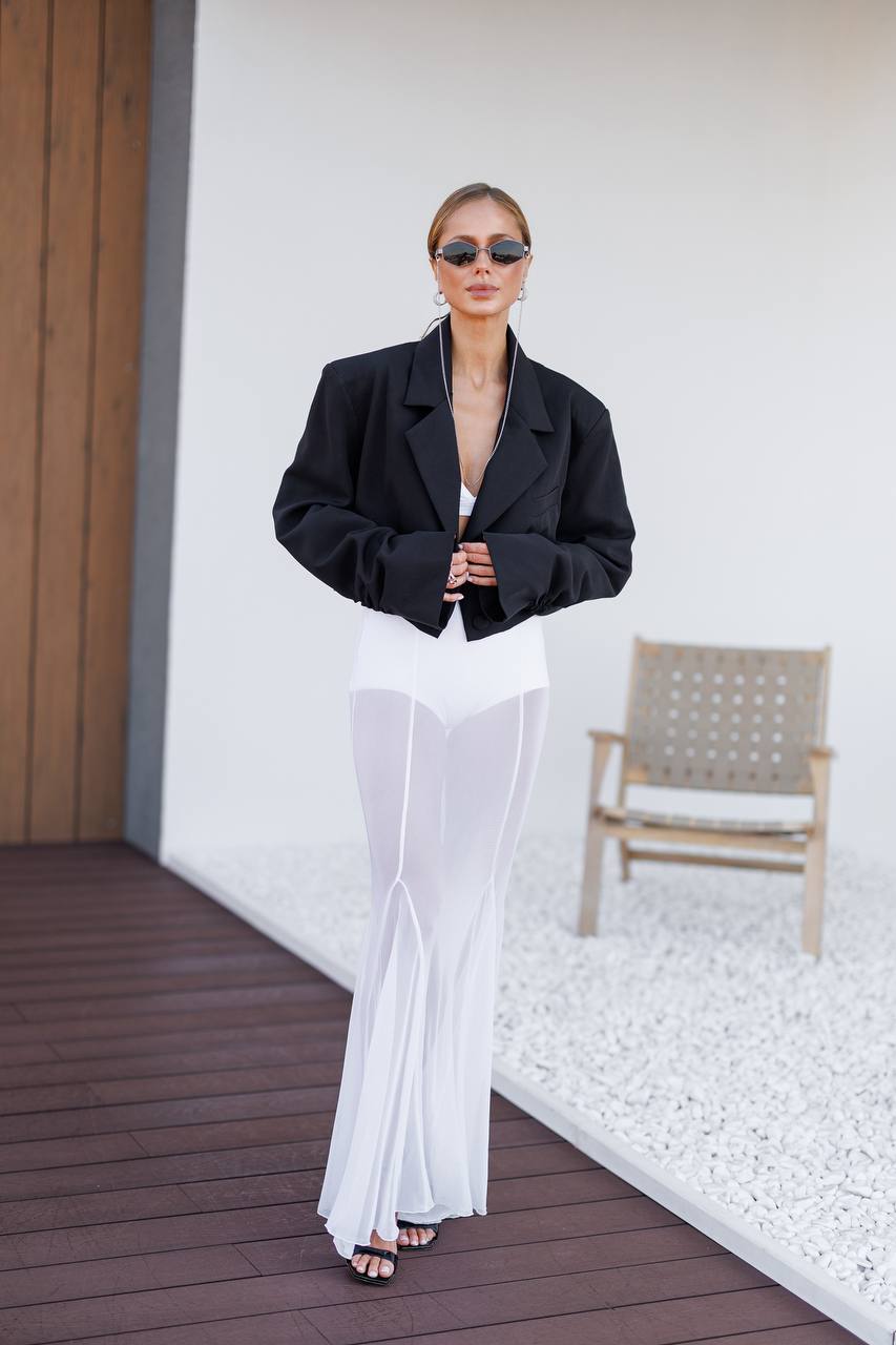 A woman stands on a wooden deck wearing a black oversized blazer, a white sheer maxi skirt, and black sunglasses. A white wall, a woven chair, and white gravel are in the background.