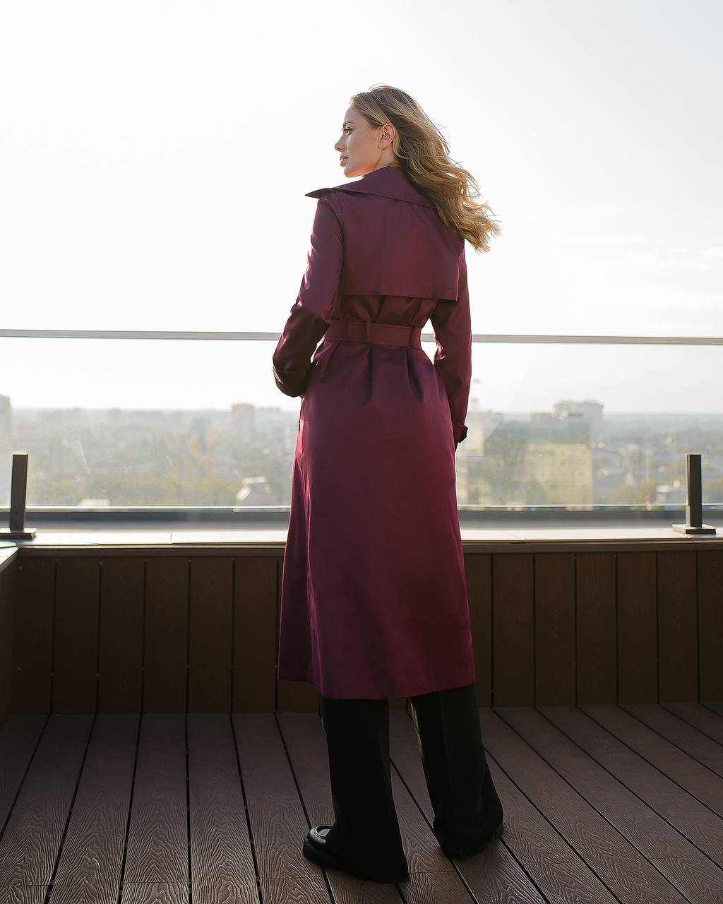 Woman in a long pink coat standing on a balcony with a cityscape view.