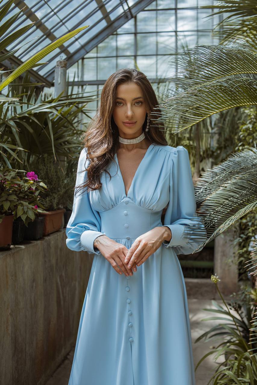 Woman in a light blue dress standing in a greenhouse filled with plants.