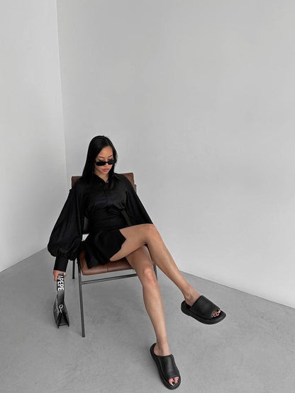 Person wearing a black outfit sitting on a chair against a plain background
