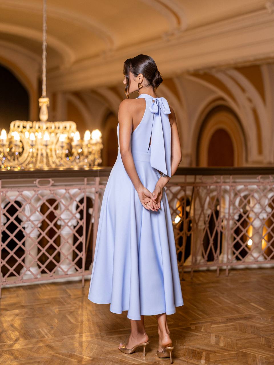 Woman in a light blue dress with a bow detail standing in an elegant interior setting.