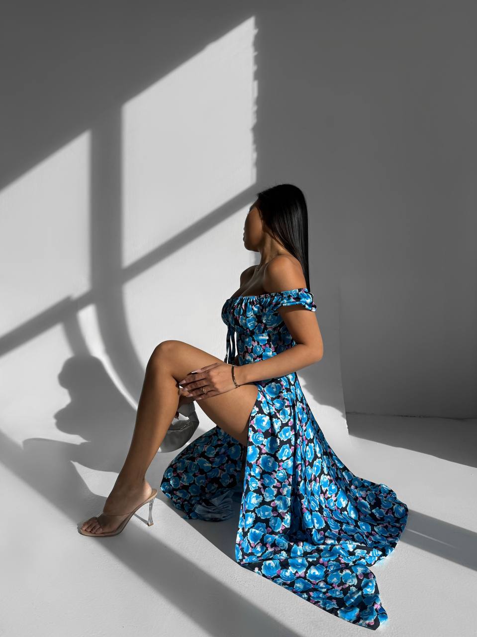 Woman in a blue floral dress sitting against a gray wall with light casting shadows.