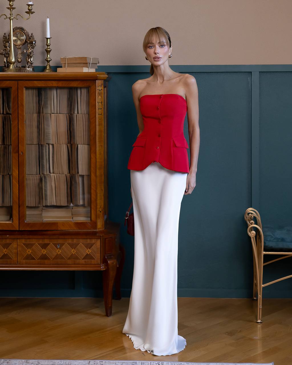 A woman stands indoors wearing a strapless red peplum-style top and a long white skirt. The room has a wood cabinet, dark teal wall paneling, and a wooden floor.