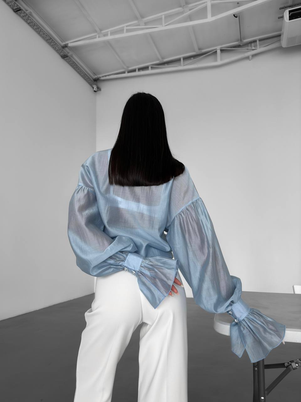 Person wearing a light blue sheer blouse with white pants in a minimalistic room.