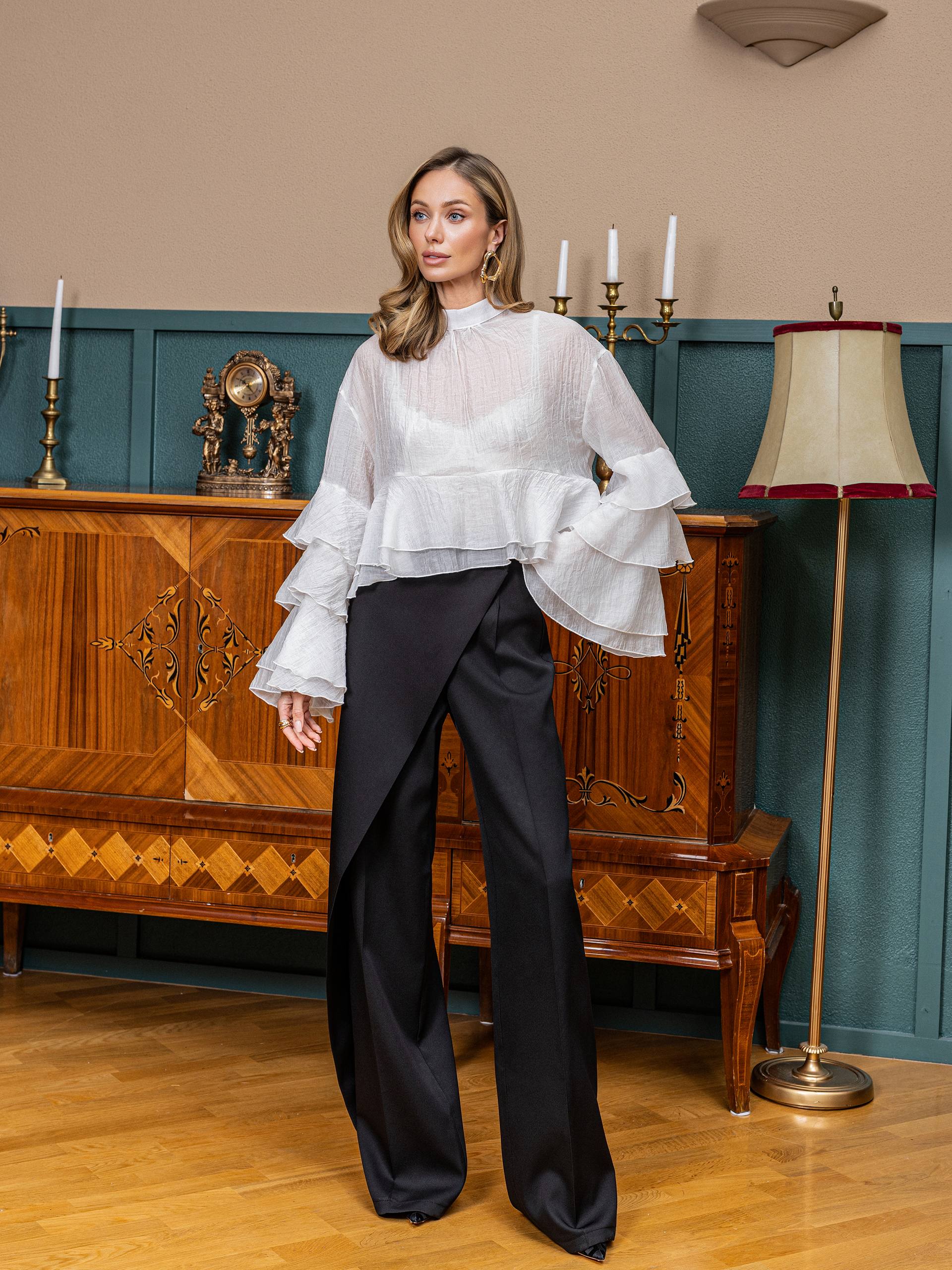 Woman in a white blouse and black pants standing in a room with wooden furniture and decorative elements.