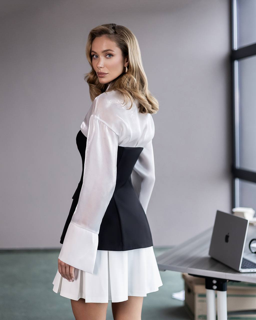 A woman models a color-blocked mini dress with a sheer white long-sleeve top overlay, a black corset-style midsection, and a white pleated skirt, set in a modern office environment.