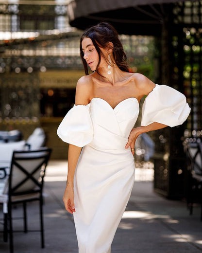 Woman in a white off-shoulder dress with puffed sleeves standing outdoors.